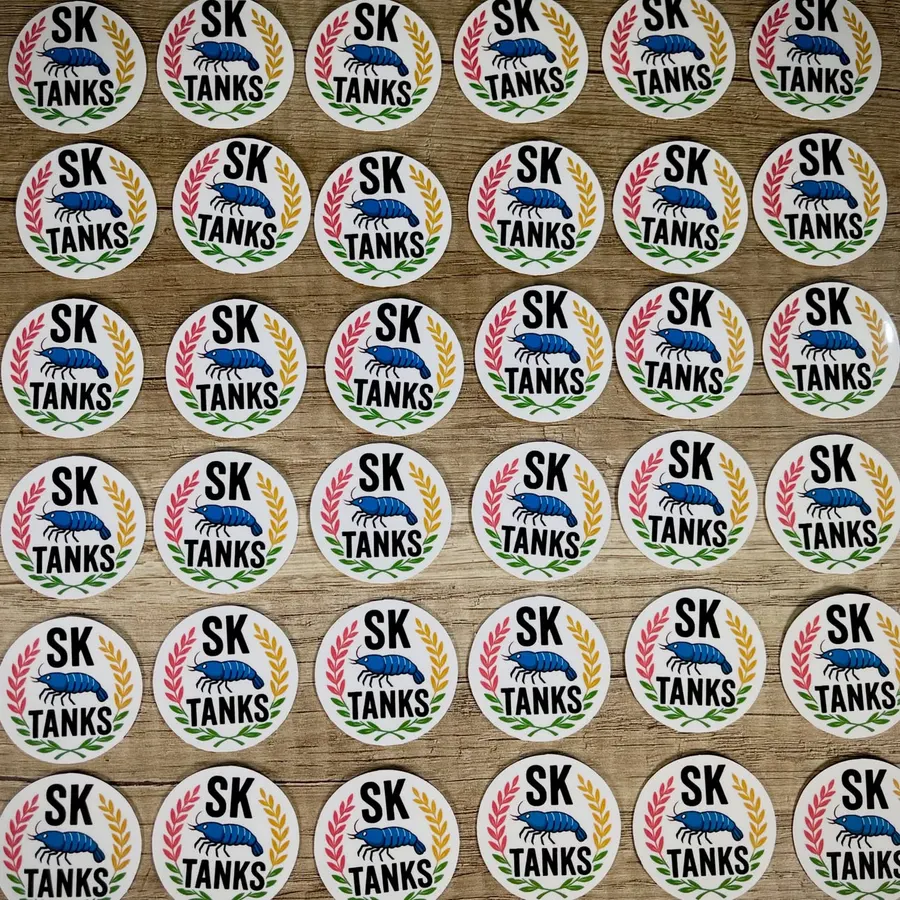 Viele runde Aufkleber mit dem SK-Tanks-Logo und Garnelenmotiv, auf einer Holzplatte ausgelegt